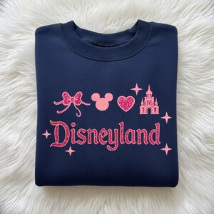 May include: Navy blue sweatshirt with pink glitter Disneyland text and graphics, including a bow, Mickey Mouse head, heart, castle, and stars. The sweatshirt is a crew neck style.