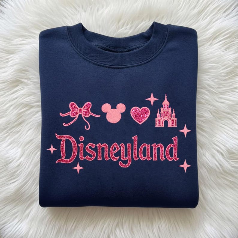 May include: Navy blue sweatshirt with pink glitter Disneyland text and graphics, including a bow, Mickey Mouse head, heart, castle, and stars. The sweatshirt is a crew neck style.