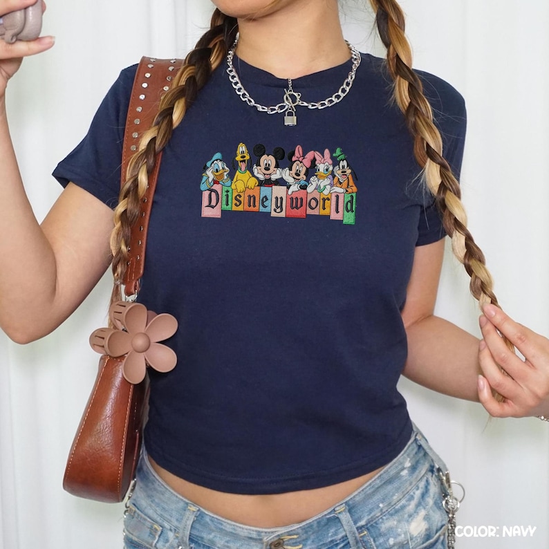 May include: A navy blue t-shirt with an embroidered Disney characters design and the text "Disneyworld". The top has short sleeves and a crew neck. A brown handbag with a flower clasp is also visible.