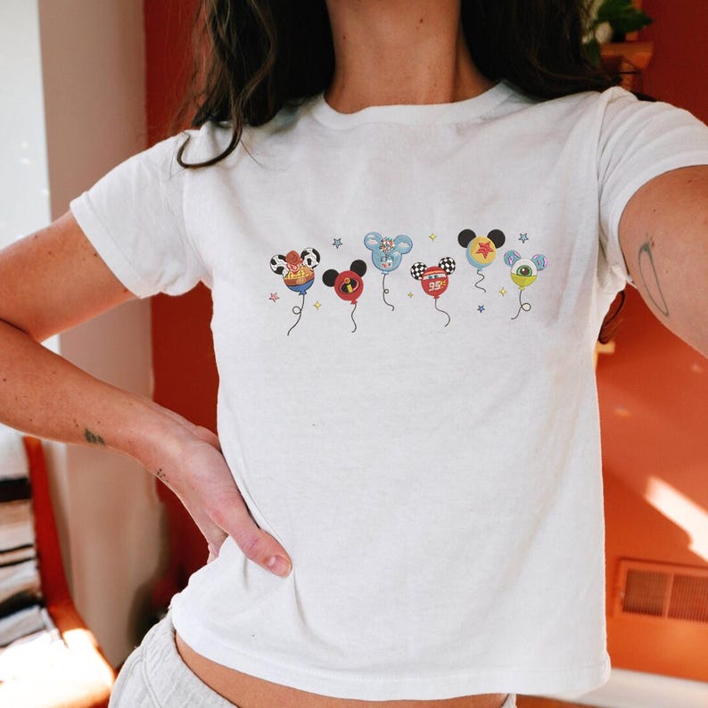 May include: White t-shirt featuring a series of cartoon character-themed balloon designs. The balloons are in the shape of Mickey Mouse heads, each with a unique design and color scheme. The shirt is short-sleeved and appears to be made of cotton.