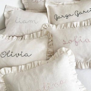 Könnte beinhalten: Eine Sammlung beiger Leinen-Zierkissen mit Rüschenkanten. Jedes Kissen hat gestickte Namen in verschiedenen Farben, darunter Schwarz und Rosa. Die Namen sind "Liam", "Olivia", "Sofia" und "Garza Garcia".