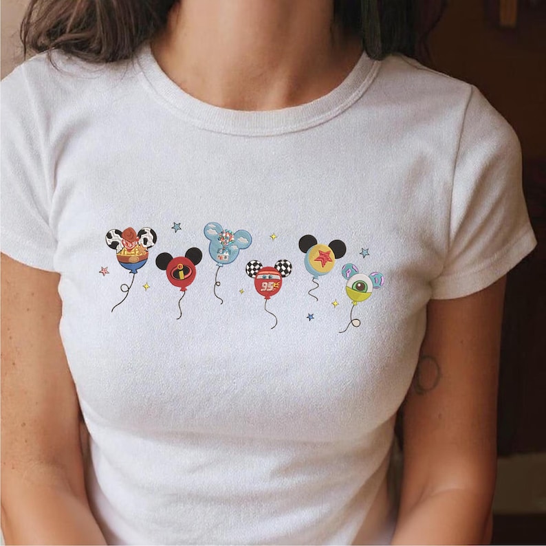 May include: White t-shirt featuring colorful cartoon character balloon designs. The balloons include a cowboy hat, a red and black balloon, a blue balloon, a checkered balloon with "95" on it, a yellow star balloon, and a green eye balloon.
