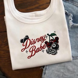 Peut inclure: Débardeur blanc côtelé avec l'inscription "Disney Babe" en rouge, accompagné de graphiques de cerises et de dés. Les cerises ont des oreilles de Mickey Mouse. Le débardeur est plié.
