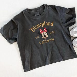 May include: Dark gray t-shirt with the Disneyland logo embroidered in gold thread. The design includes Minnie Mouse with a red and white polka dot bow, the text "est. 1955 California". The shirt has short sleeves and a crew neck.