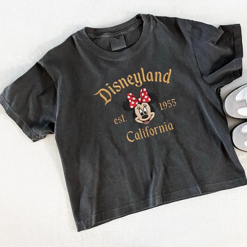 May include: Dark gray t-shirt with the Disneyland logo embroidered in gold thread. The design includes Minnie Mouse with a red and white polka dot bow, the text "est. 1955 California". The shirt has short sleeves and a crew neck.