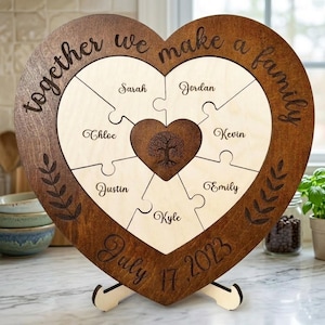 May include: A heart-shaped wooden puzzle plaque featuring the engraved text "together we make a family" and "July 17, 2023". The puzzle contains six light-coloured pieces with names and a central dark heart piece with a tree illustration.