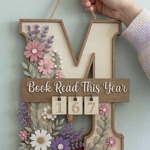 May include: A wooden wall hanging shaped like the letter M, featuring floral cutouts in shades of pink, purple, and white. A horizontal bar displays the text "Book Read This Year" above three small, interchangeable wooden tiles showing the number 167.