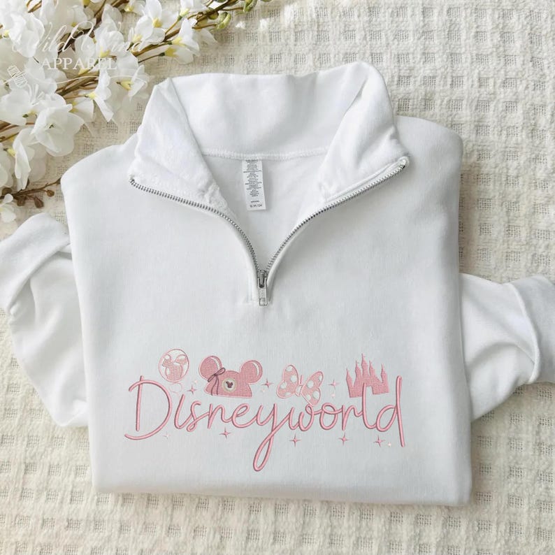 May include: White quarter-zip sweatshirt with a pink Disneyworld design. The design includes a balloon, Minnie Mouse head, bow, and castle. The text "Disneyworld" is in a cursive font.