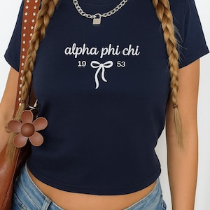 Può includere: Top corto blu navy con la scritta bianca "alpha phi chi", i numeri "19 53" e un fiocco. Indossata una collana a catena argentata con un ciondolo a lucchetto. La persona indossa jeans blu e ha lunghi capelli intrecciati.