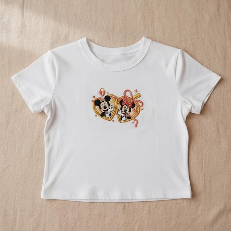 May include: White crop top featuring an embroidered design of Mickey and Minnie Mouse inside heart-shaped frames. The design includes gold, red, and black colors. The shirt has short sleeves and a crew neck.
