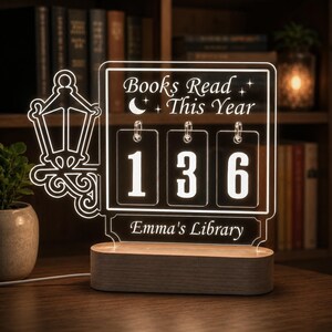 Könnte beinhalten: Ein durchsichtiges Acryl-Schild zur Buchverfolgung mit Holzsockel, das den Text "Books Read This Year" und "Emma's Library", eine Laternen-Grafik sowie drei hängende rechteckige Acryl-Schilder mit den weißen Ziffern 1, 3 und 6 zeigt.
