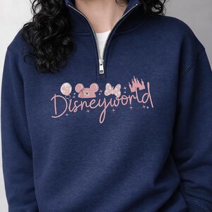 May include: Navy blue sweatshirt with a half-zip collar. The front features a pink graphic of Mickey Mouse, a bow, a castle, and the word "Disneyworld" in cursive. The sweatshirt has a white undershirt peeking out.