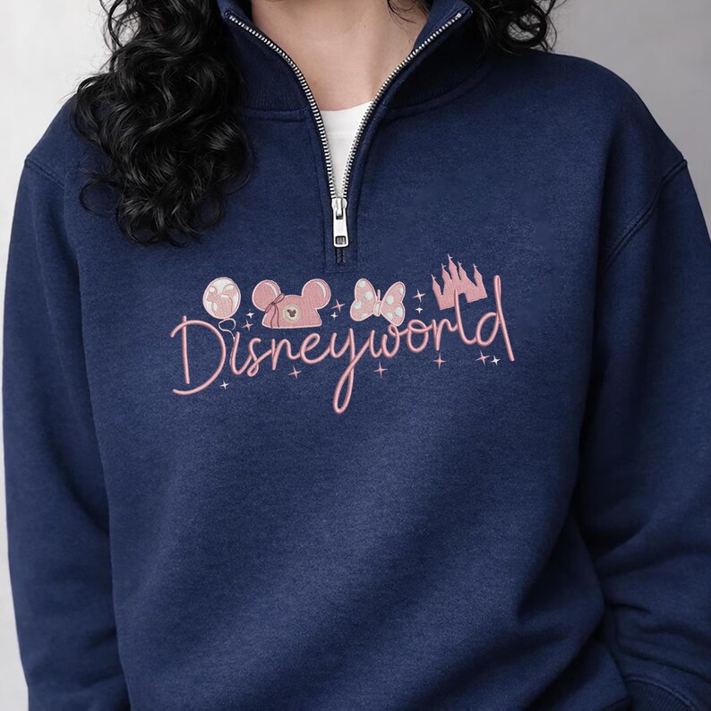 May include: Navy blue sweatshirt with a half-zip collar. The front features a pink graphic of Mickey Mouse, a bow, a castle, and the word "Disneyworld" in cursive. The sweatshirt has a white undershirt peeking out.