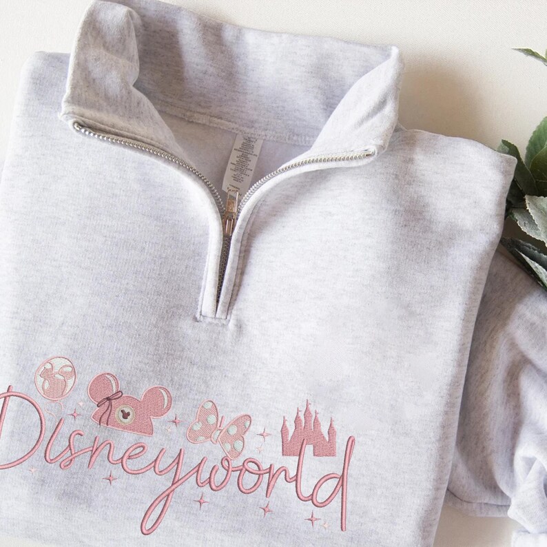 May include: A light gray quarter-zip sweatshirt with a pink embroidered "Disneyworld" design. The design includes a Mickey Mouse head, a castle, and other Disney-themed elements. The sweatshirt has a high collar and a silver zipper.