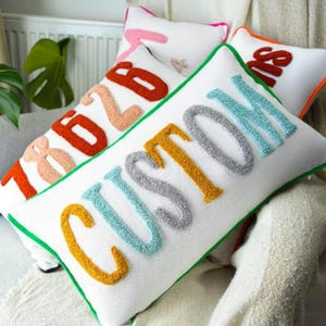 May include: White rectangular pillow with the word "CUSTOM" in yellow, gray, and blue textured letters. Other pillows in the background have numbers and words in orange, pink, and red. The pillows have green and orange trim.