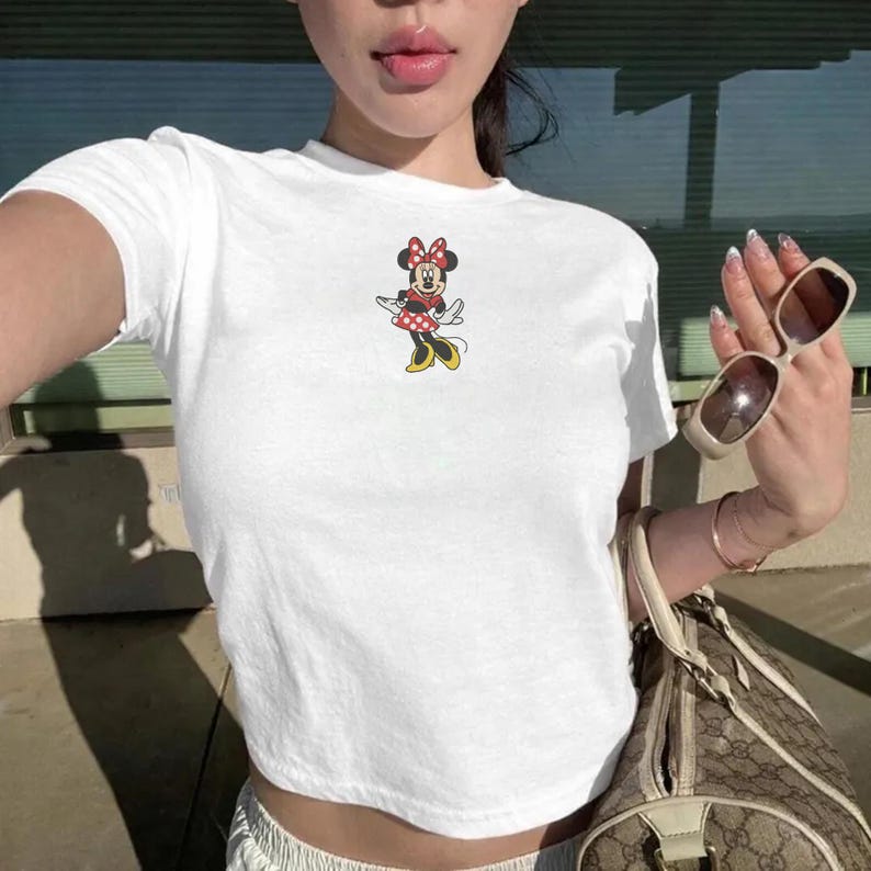 Puede incluir: Top corto blanco con un gr&aacute;fico de Minnie Mouse. El gr&aacute;fico muestra a Minnie Mouse con un vestido de lunares rojos, un lazo y zapatos amarillos. La parte superior est&aacute; hecha de un material suave y c&oacute;modo.