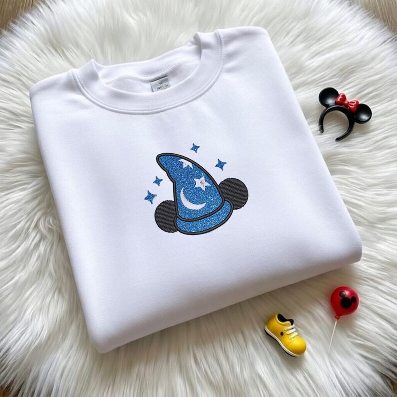 Op de afbeelding: Witte sweatshirt met een blauwe glitter tovenaars hoed en Mickey Mouse oren ontwerp. De hoed heeft een maan en sterren. Een Minnie Mouse oor hoofdband, een gele schoen en een Mickey Mouse ballon staan ook in de afbeelding.