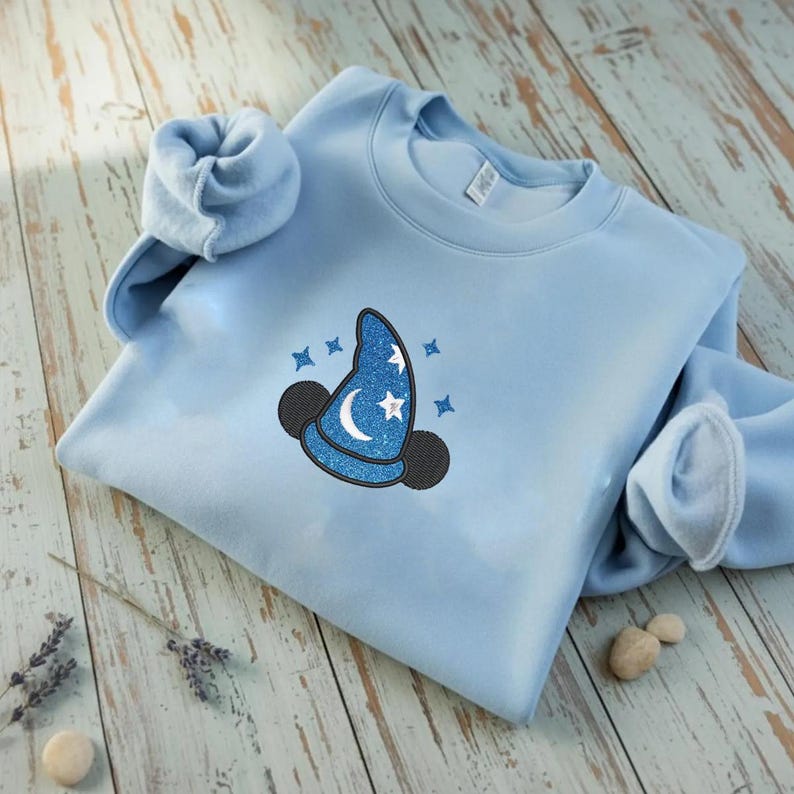 Op de afbeelding: Een lichtblauw sweatshirt met een grillig ontwerp. Het ontwerp omvat een blauwe tovenaars hoed met glitter, een maan, sterren en zwarte Mickey Mouse oren. Het sweatshirt is op een licht houten oppervlak gevouwen.