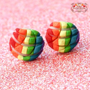 May include: A pair of colorful stud earrings. Each earring is a sphere with a rainbow pattern of red, orange, yellow, green, blue, and purple. The earrings are on a pink glitter surface. The earrings are handmade.