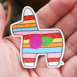 May include: A colorful pinata shaped like a donkey with a pink flower and green leaf on its side. The pinata is decorated with stripes of red, orange, yellow, blue, and pink.