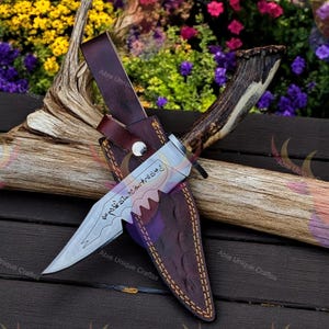 Può includere: Un coltello da caccia con una lama argentata e un manico in corno marrone, in un fodero di cuoio marrone. Il fodero è cucito con un motivo decorativo. La lama ha un testo inciso. Il coltello è esposto su legno di deriva.