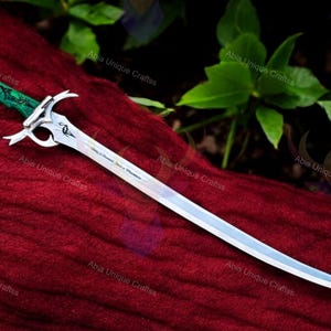 May include: A decorative sword with a curved, silver blade and a green handle. The handle has a detailed design, and the blade has an inscription. The sword rests on a red fabric, with green foliage in the background.