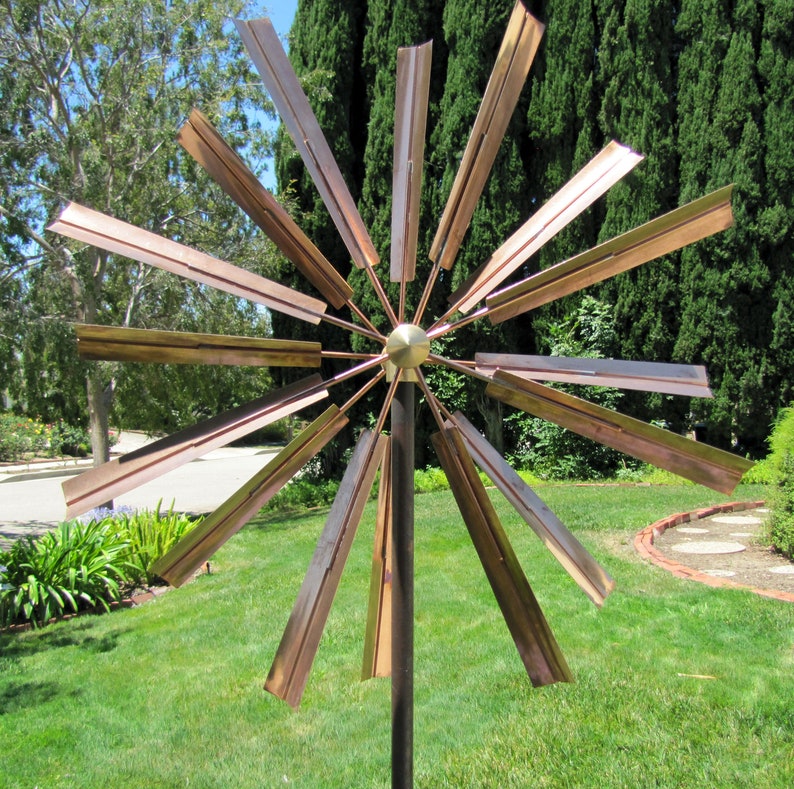 May include: A copper-colored metal wind spinner with 12 blades, mounted on a black metal pole. The blades are arranged in a starburst pattern and are designed to spin in the wind.
