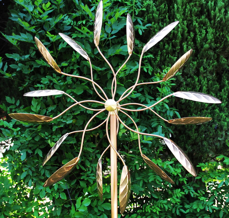 May include: A gold metal wind spinner with multiple leaf-shaped blades. The spinner is mounted on a tall pole and is surrounded by green foliage.