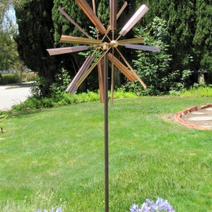 May include: A copper and gold metal wind spinner with multiple blades, mounted on a tall metal pole. The spinner is in a garden setting with green grass and purple flowers.