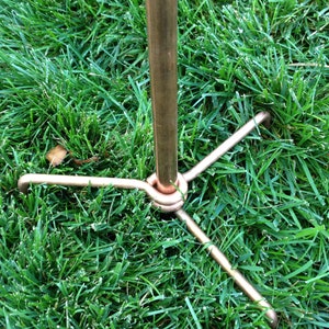 May include: A copper metal stand with three legs. The stand is designed to be inserted into the ground.
