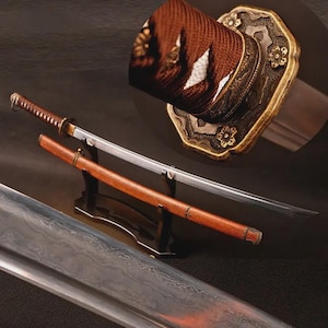 May include: A katana sword with a curved steel blade featuring a visible wavy temper line, a brown cord-wrapped handle, and a wooden scabbard. The sword is displayed on a black stand, with a detailed close-up of the ornate gold-toned metal handguard.