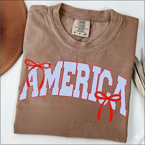 Pode incluir: Uma camiseta Comfort Colors marrom com a palavra "AMERICA" em letras brancas com listras azuis, contornada em vermelho. Laços vermelhos estão nas letras. Um chapéu marrom é visível no canto superior esquerdo.