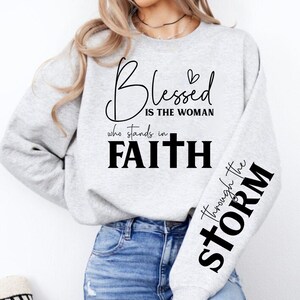 May include: A light gray sweatshirt with black script that reads "Blessed is the woman who stands in Faith" on the front. The right sleeve has the words "through the STORM" in black. The sweatshirt is a crew neck style.