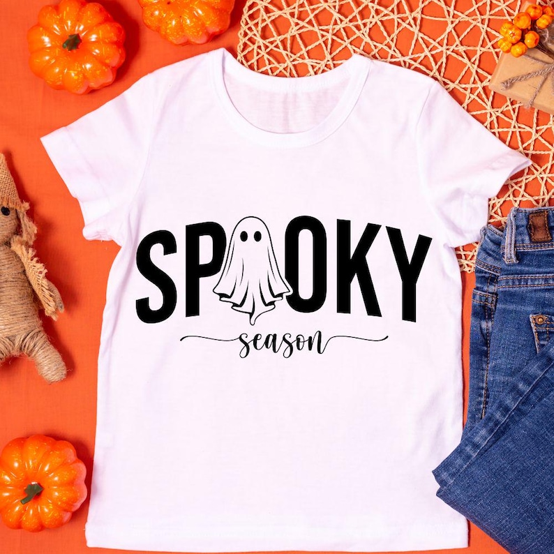 K&ouml;nnte beinhalten: Wei&szlig;es T-Shirt mit den schwarzen Worten "SPOOKY season" und einer Geistergrafik. Das Shirt wird auf einer orangefarbenen Oberfl&auml;che mit K&uuml;rbissen und Jeans im Hintergrund pr&auml;sentiert. Ein festliches Halloween-Thema.