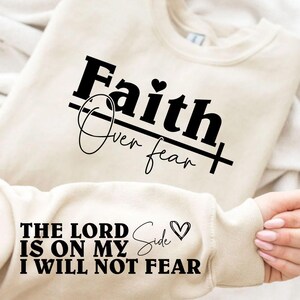 K&ouml;nnte beinhalten: Cremefarbenes Sweatshirt mit dem Text "Faith Over Fear" in fetter, schwarzer Schrift, mit einem Herzsymbol. Der &Auml;rmel zeigt den Satz "The Lord Is On My Side, I Will Not Fear". Ein l&auml;ssiges, inspirierendes Kleidungsst&uuml;ck.