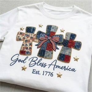 Può includere: Una t-shirt bianca con tre applicazioni a forma di croce in un motivo patchwork di tessuto rosso, bianco e blu con motivi di bandiere americane. Un fiocco a righe si trova sulla croce centrale. Il testo recita God Bless America Est. 1776 con stelle dorate.