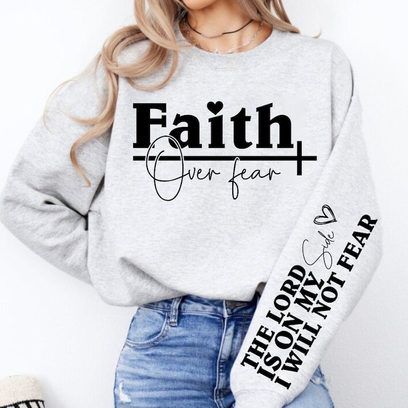 K&ouml;nnte beinhalten: Hellgraues Sweatshirt mit dem Schriftzug "Faith Over Fear" in schwarzen Buchstaben. Der &Auml;rmel zeigt den Text "The Lord is on my side, I will not fear" mit einem Herzsymbol. Ein l&auml;ssiges, inspirierendes Design.