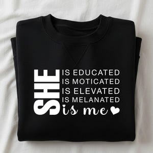 May include: Black sweatshirt with white text that reads "SHE IS EDUCATED IS MOTIVATED IS ELEVATED IS MELANATED is me" with a heart symbol. The sweatshirt is folded neatly on a white surface.