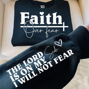 K&ouml;nnte beinhalten: Schwarzes Sweatshirt mit dem wei&szlig;en Schriftzug "Faith Over Fear". Der &Auml;rmel zeigt "The Lord Is On My Side I Will Not Fear" in Wei&szlig;. Ein Herz-Grafik ist im Design enthalten.