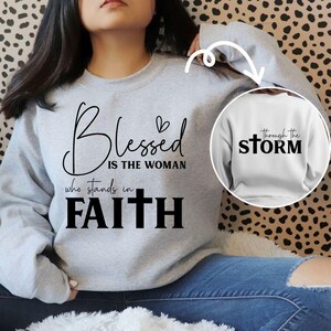 May include: A light gray sweatshirt with the words "Blessed is the woman who stands in Faith" in black script. A smaller image shows a white sweatshirt with the words "through the STORM". The sweatshirt is a casual, comfortable style.