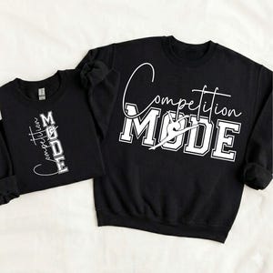 Puede incluir: Dos sudaderas negras con texto blanco. La sudadera más grande tiene las palabras "Competition MODE" con una silueta de gimnasta. La sudadera más pequeña tiene las palabras "Competition MODE" verticalmente. Ambas sudaderas están sobre un fondo blanco.