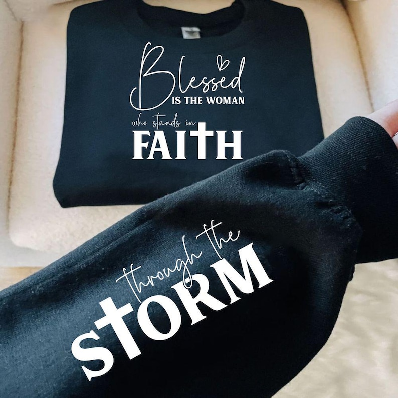 May include: Black sweatshirt with the text "Blessed is the woman who stands in Faith" in white script. The sleeve displays the phrase "Through the Storm" in white script. The sweatshirt is displayed on a cream-colored surface.