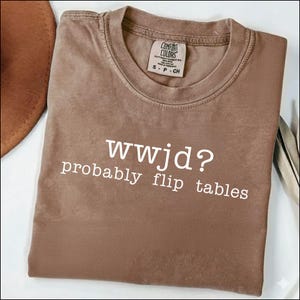 May include: A brown t-shirt with the text "wwjd? probably flip tables" in white. The shirt has a crew neck and a small tag on the collar. The shirt is folded and appears to be made of a soft material.