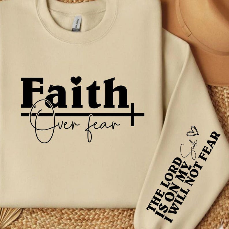 K&ouml;nnte beinhalten: Ein cremefarbenes Sweatshirt mit den Worten "Faith Over Fear" in fetter schwarzer Schrift. Der &Auml;rmel zeigt den Text "The Lord is on my side, I will not fear" in Schwarz. Ein Herzsymbol ist enthalten.