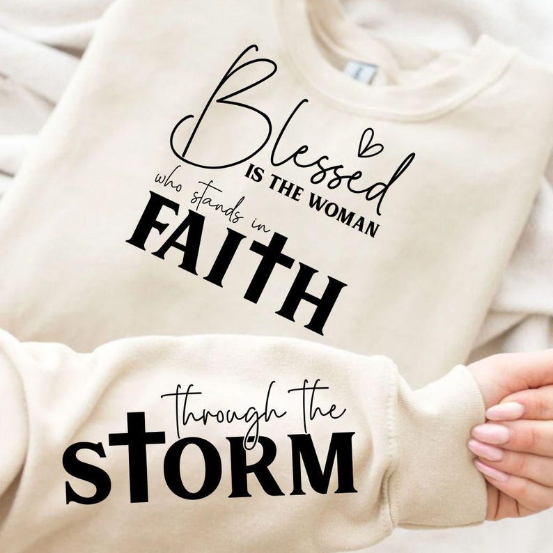 May include: Cream-colored sweatshirt with the words "Blessed is the woman who stands in Faith" in black script. The sleeve displays "Through the Storm" in black lettering. The sweatshirt is made of a soft material.