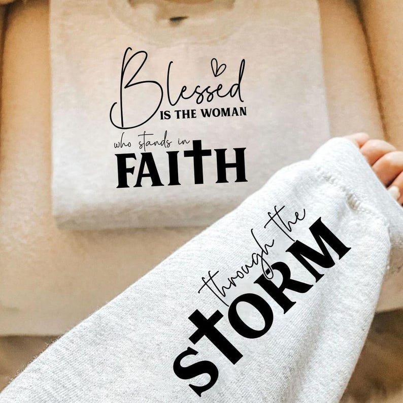 May include: A folded light gray sweatshirt with the words "Blessed is the woman who stands in Faith" printed in black. A sleeve with the words "through the STORM" is visible.