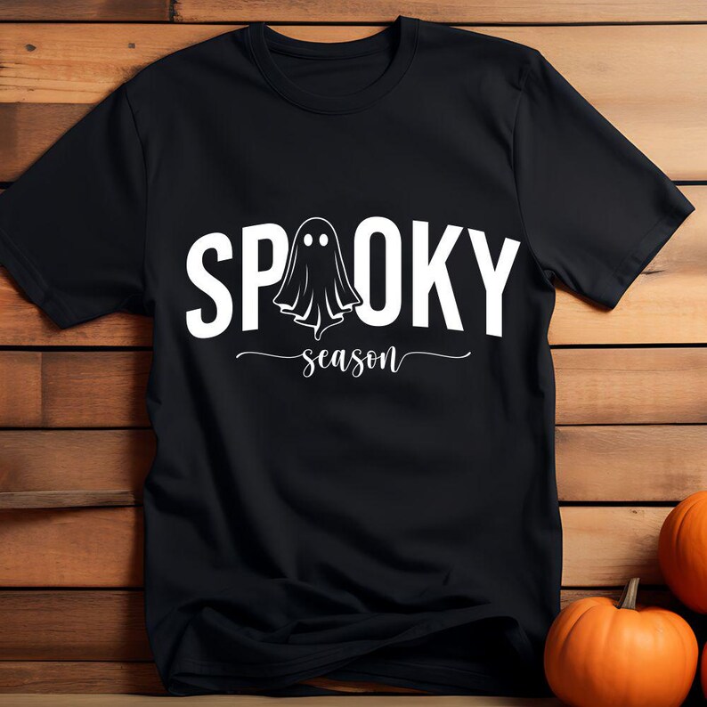 K&ouml;nnte beinhalten: Schwarzes T-Shirt mit dem wei&szlig;en Schriftzug "SPOOKY season" und einer Geistergrafik. Das Shirt wird auf einer Holzoberfl&auml;che mit zwei orangefarbenen K&uuml;rbissen im Hintergrund pr&auml;sentiert, perfekt f&uuml;r Halloween.
