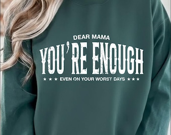 May include: A dark teal sweatshirt with the text "DEAR MAMA YOU'RE ENOUGH" in white, bold, capital letters. Below the main text, the phrase "EVEN ON YOUR WORST DAYS" is written in smaller white letters, with three white stars on each side.