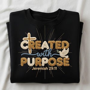 Op de afbeelding: Zwart sweatshirt met de tekst "CREATED with PURPOSE" in een gehaakte lettertype, een kruis en een duif met een olijftak. De tekst "Jeremiah 29:11" staat eronder. Het ontwerp gebruikt goud- en crèmekleuren.