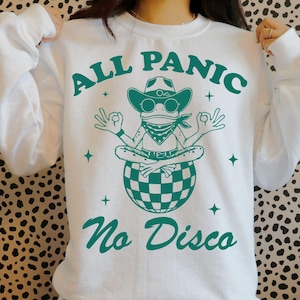 Könnte beinhalten: Weißes Sweatshirt mit dem Text "ALL PANIC No Disco" in Türkis. Die Grafik zeigt einen Frosch mit Cowboyhut, Sonnenbrille und Halstuch, der auf einer karierten Discokugel sitzt und die Hände in einer Yoga-Pose hält.